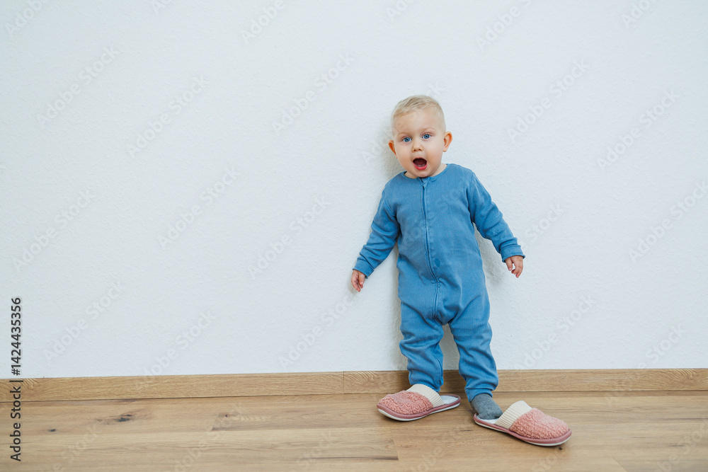Little toddler boy in a blue romper tries on big house slippers. Child plays at home in the living room. Children's home clothes.