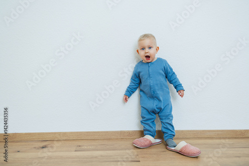 Little toddler boy in a blue romper tries on big house slippers. Child plays at home in the living room. Children's home clothes.