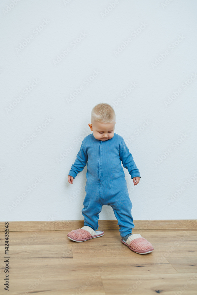 Little toddler boy in a blue romper tries on big house slippers. Child plays at home in the living room. Children's home clothes.