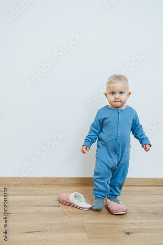 Little toddler boy in a blue romper tries on big house slippers. Child plays at home in the living room. Children's home clothes.