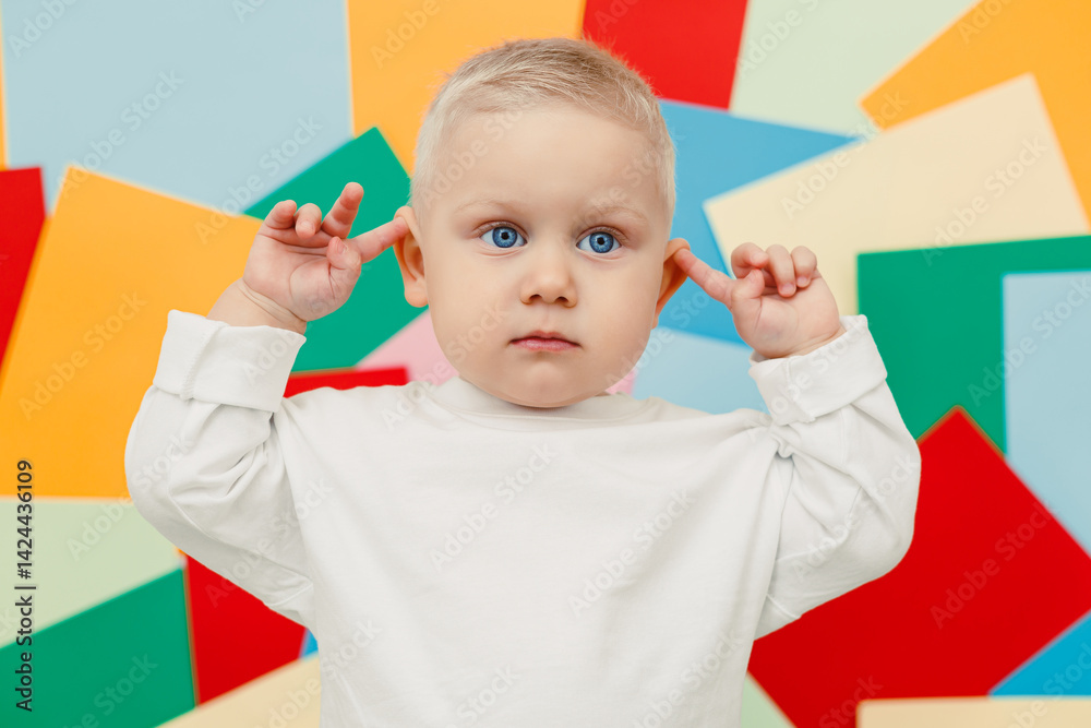 Happy little toddler in a white t-shirt and jeans stands on a studio background and covers his ears, expressing joy and curiosity. Stylish toddler. Concept of parenthood, natural beauty, love and care
