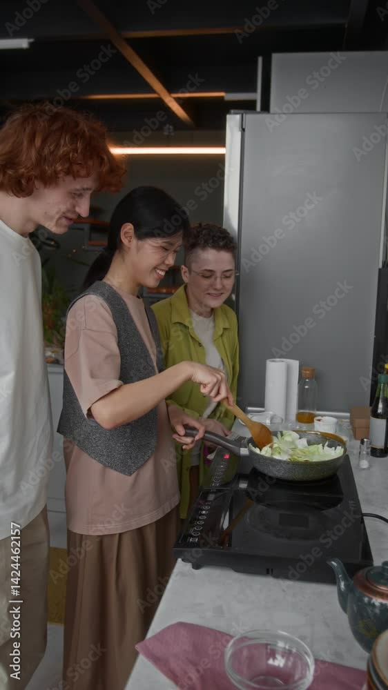 Vertical view of three multiethnic young friends watching Asian girl who cooking Chinese dish, Caucasian man taking cabbage leaf from frying pan Vertical view of three multiethnic young friends watching Asian girl who cooking Chinese dish, Caucasian man taking cabbage leaf from frying pan