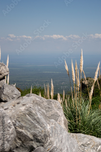 Dramatic vista from a mountain edge with dry grass and rocks in the foreground. A clear sky and distant valley create a sense of scale and solitude. A peaceful and expansive mountain moment.