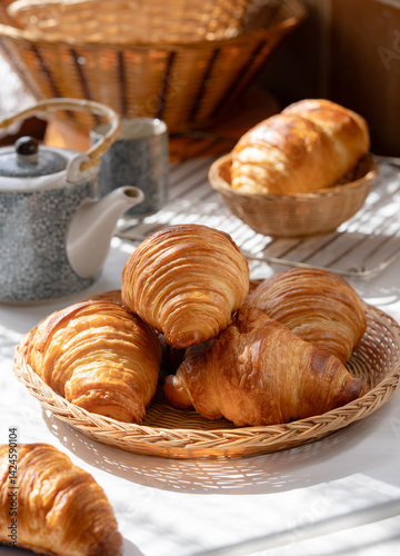 Homemade French butter croissant in wooden basket with cup and pot of tea, cozy ambiance of sunlight of the morning from window shade.