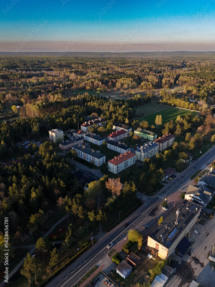 Fototapeta premium Aerial view of Stąporków, Poland, with apartment buildings nestled among forested areas, showcasing the balance of urban life and nature during golden hour.