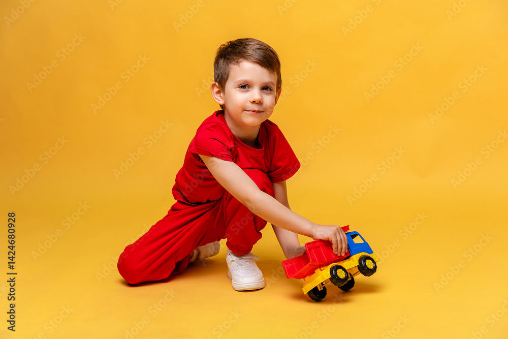 Child, little boy in red sport suit playing with toy car on yellow background. Childhood education, kindergarten and development concept, Space for text. High quality photo.