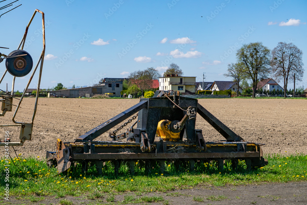 Fototapeta premium An agricultural machine stands on the edge of a field in a rural landscape. Single-family homes and trees are visible in the background.
