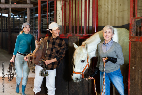 Obraz na płótnie Positive Asian horsewoman holding saddle while elderly female stable keeper leading white horse out of stall by bridle.. w salonie
