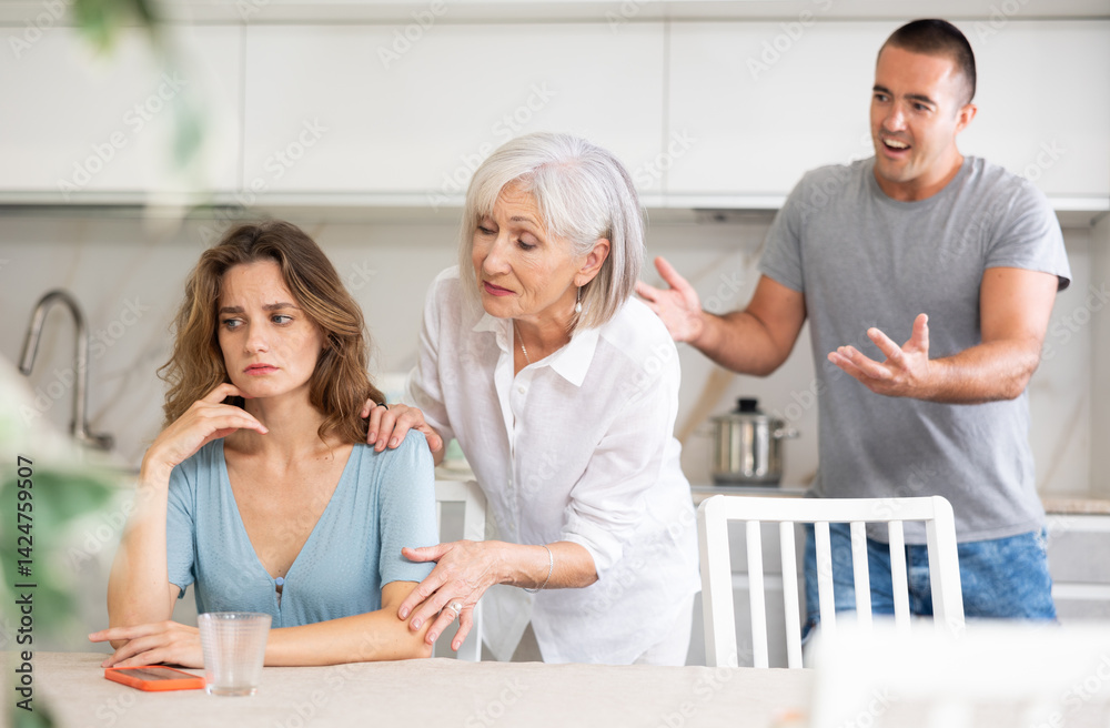 Fototapeta premium Young girl is sitting at table in kitchen quarreling with her mother-in-law and husband