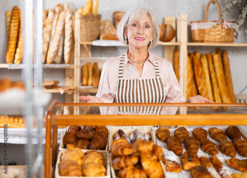 Obraz na płótnie Portrait of smiling elderly woman baker standing behind display case with fresh croissants. Offering to buy fresh baked goods w salonie