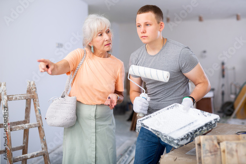 Senior woman employer explaining work details to teenager boy who working on part-time job in construction site as painter. Woman making pointing finger gesture.