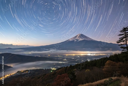 A wide-angle view of Mount Fuji surrounded by a mountainous landscape, under a sky filled with star trails that create the illusion of a rotating galaxy. The scene blends twilight tones with cosmic wo