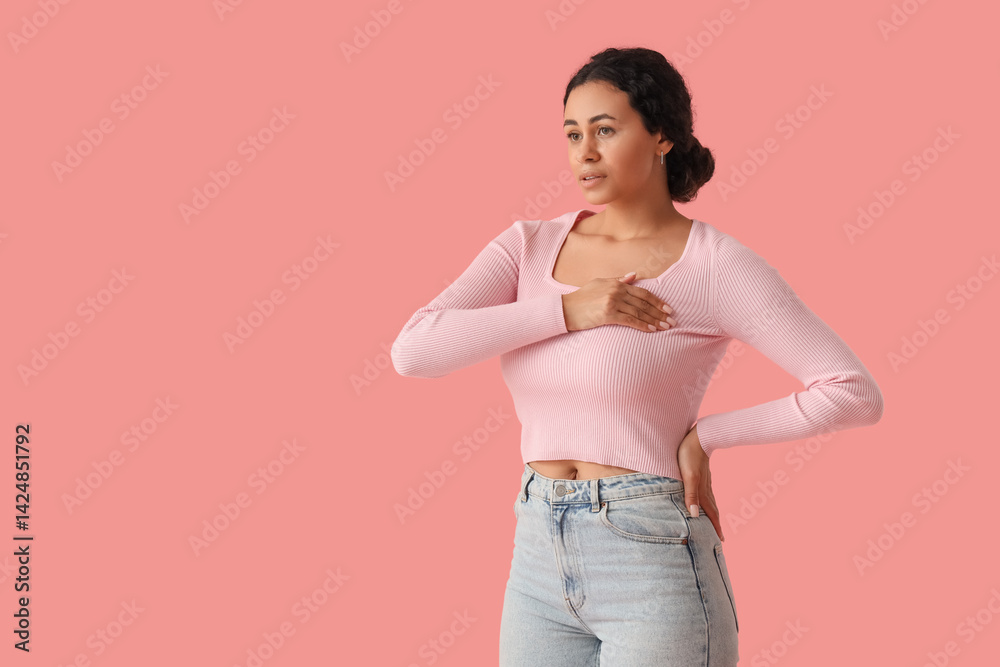 Young African-American woman checking her breast on pink background. Cancer awareness concept