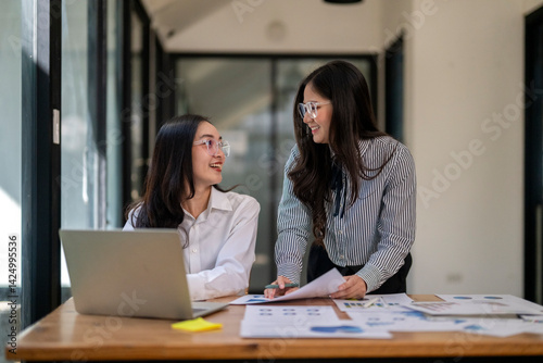 Two women are sitting at a desk with a laptop and papers in front of them