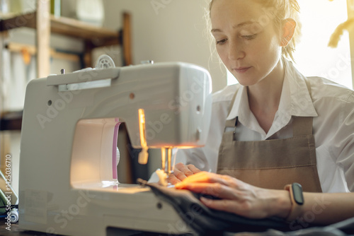 A female tailor is working on a sewing machine stitching fabric in sunny room. Home sewing production