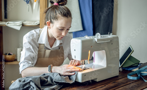 A female tailor is working on a sewing machine stitching fabric in sunny room. Home sewing production