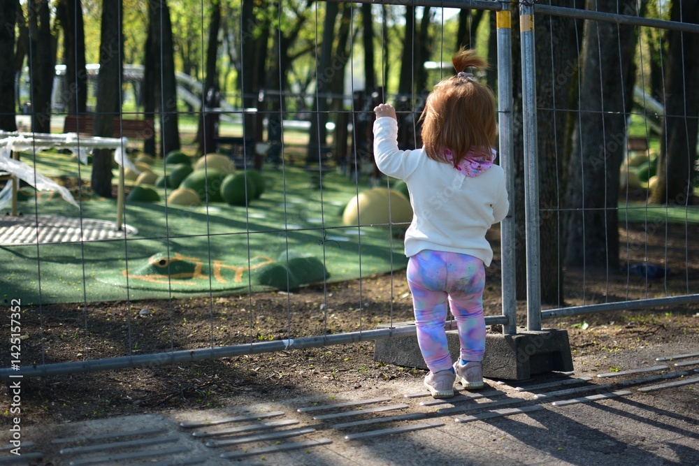 Curious red hair girl standing backwards and observing children playground construction site in spring. Toddler standing near closed playground fence waiting to play. Curious red hair girl standing backwards and observing children playground construction site in spring. Toddler standing near closed playground fence waiting to play.