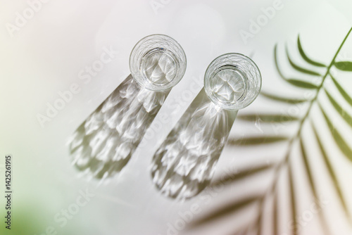 Obraz na płótnie Water Glasses with palm leaf Shadow and sunlight Reflection on white background, colored photo minimal style, aesthetic top view summer still life, refraction throug crystal shine cups, caustic w salonie