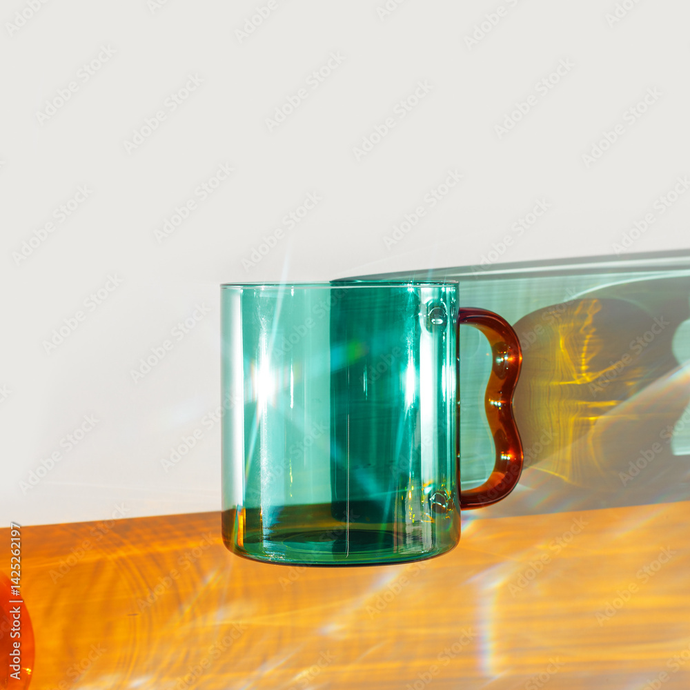 Vibrant still-life with colorful transparent glass mug or cup in shades of green teal and amber-orange. Sunlight dynamic shadows and reflections on white, minimal aesthetic, table setting