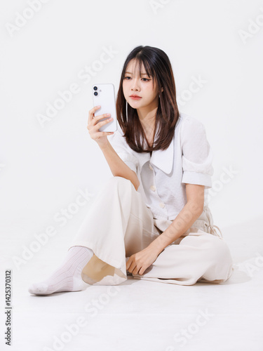 Asian young beautiful pretty happy woman she smiling, cute friendly wearing white shirt white pants sitting on floor holding smartphone on white background studio portrait. Lifestyle fashion concept.