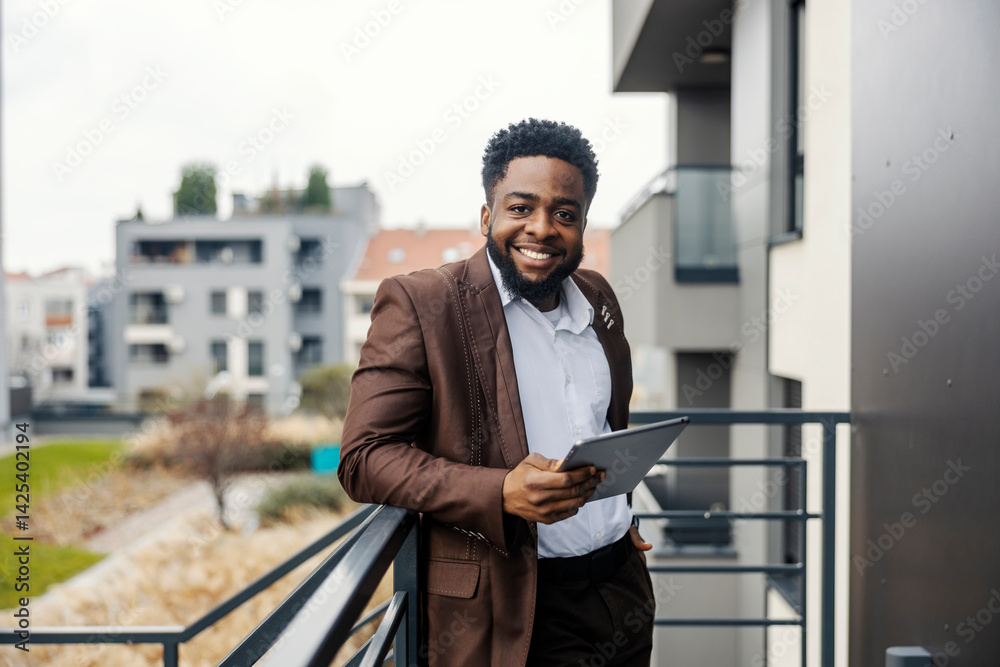 Portrait of smiling african american businessman leaning on railing on balcony with tablet in his hand.