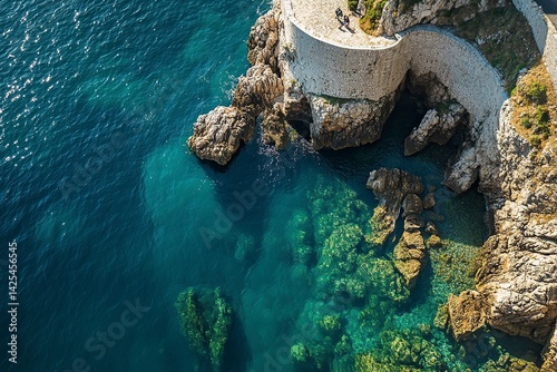 A panoramic view of the ancient city walls and sea in Dubrovnik, Croatia. The iconic Pile gate is visible on one side on a sunny summer day. Blue, clear water with rock formations in front of it. 
