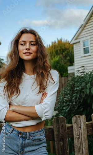 A woman with long brown hair is standing in front of a wooden fence. She is wearing a white shirt and blue jeans. Concept of casualness and relaxation