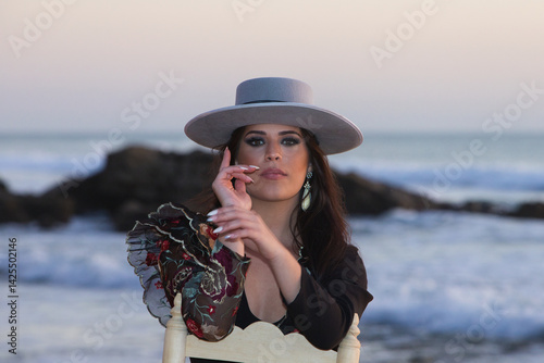 beautiful brunette spanish woman dancing flamenco with typical costume and wide brimmed hat sitting on an old wooden chair, in the background the atlantic ocean and rocks.