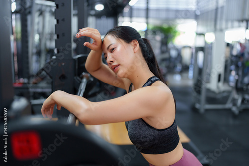 Exhausted asian sportswoman wiping sweat from her forehead while leaning on a barbell in a modern gym, demonstrating the physical demands of intense training