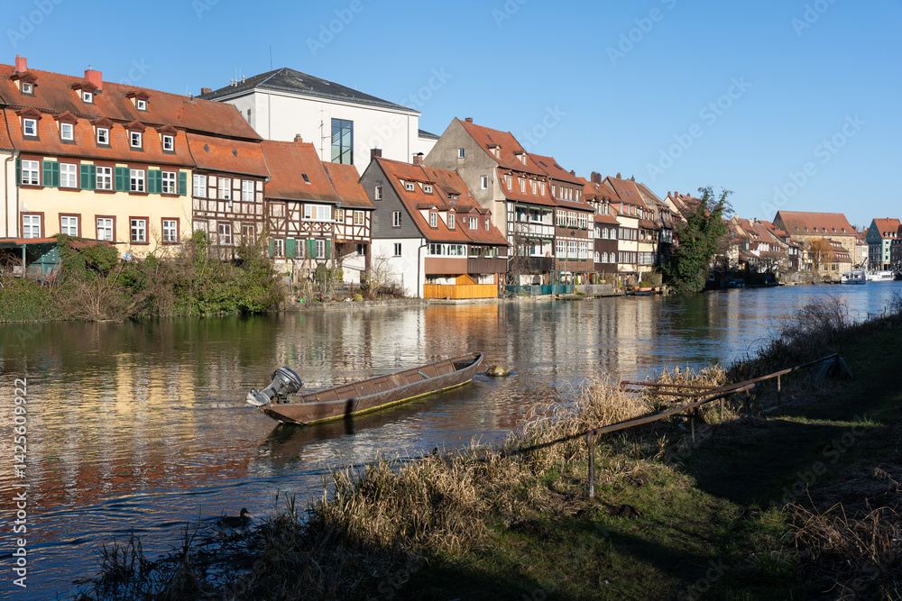 Obraz premium Old narrow motor boat in the river in German city of Bamberg with houses on the shore