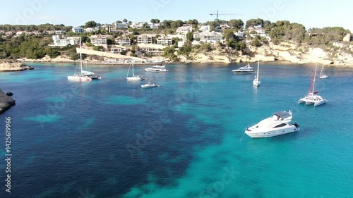 Set of several luxury yachts located in a smal bay on the coast of Mallorca.  The bay is  filled with several boats and yachts, some anchored and others moving slowly across the turquoise-blue water