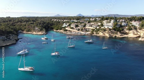 Aerial view of  luxury yachts located in a small bay on the coast of Mallorca.  The bay is  filled with several boats and yachts, some anchored and others moving slowly across the turquoise-blue water