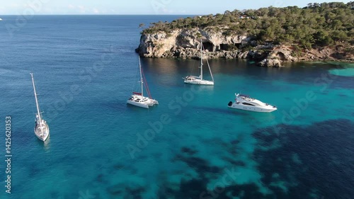  Group of several luxury yachts located in a smal bay on the coast of Mallorca.The bay is  filled with several boats and yachts, some anchored and others moving slowly across the turquoise-blue water
