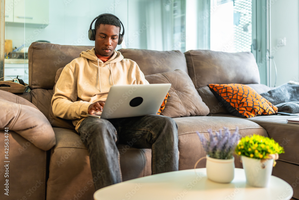 Focused teenager working on laptop with headphones on comfortable sofa at home