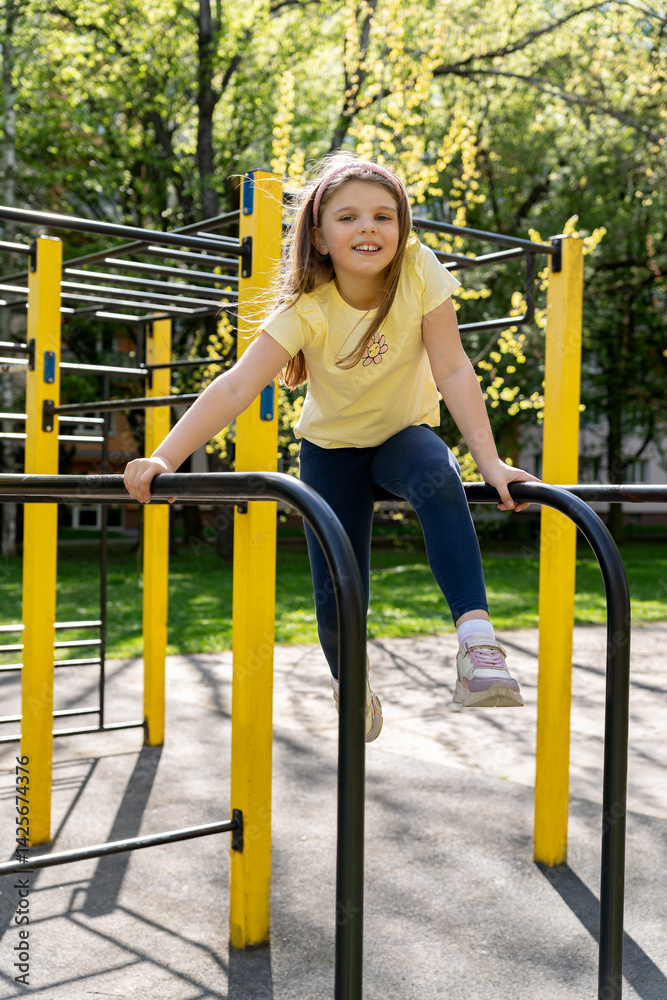 Smiling little girl in yellow t-shirt sitting on horizontal bars on school playground on sunny day. Physical education at school. Smiling little girl in yellow t-shirt sitting on horizontal bars on school playground on sunny day. Physical education at school.