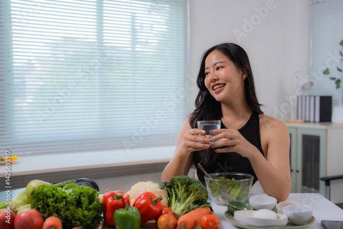 Asian nutritionist holding a glass of water and smiling while sitting near a table with fresh vegetables, promoting healthy eating habits and hydration for a balanced lifestyle