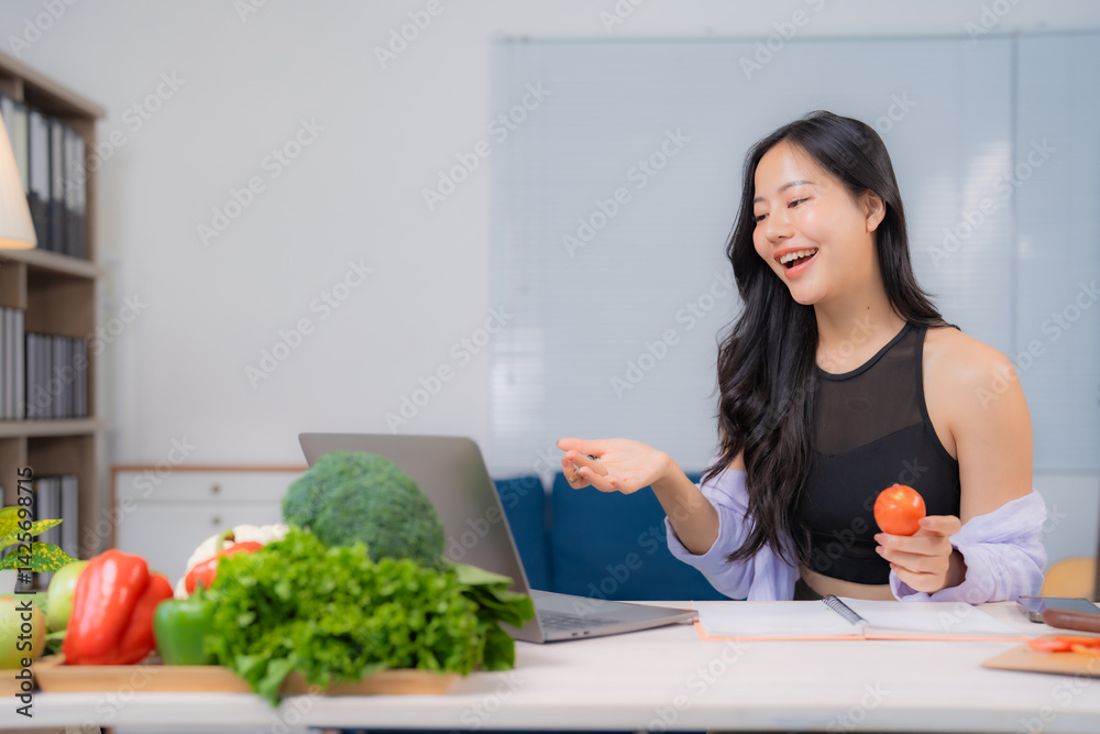 Asian nutritionist holding tomato having video call on laptop promoting healthy eating habits with vegetables and fruits lying on desk in her office Asian nutritionist holding tomato having video call on laptop promoting healthy eating habits with vegetables and fruits lying on desk in her office