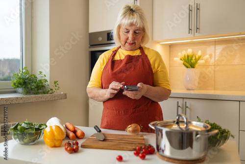 A woman in a yellow shirt and apron is preparing food in the kitchen. She looks at her phone for a recipe while chopping vegetables like tomatoes and bell peppers.