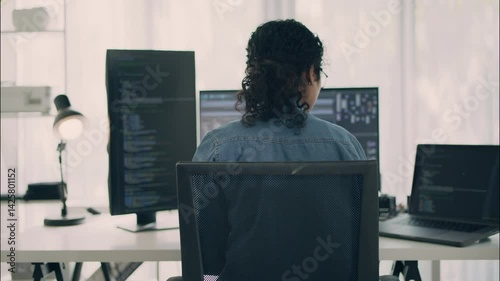 Rear view of female coder working on programming and UI development across multiple monitors in a modern tech office. Concept of multitasking and software engineering.