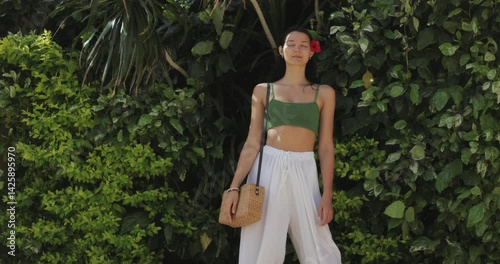 Stylish woman with long dark hair and a red flower in her hair, posing in front of lush green foliage on a windy day. She is wearing a green top and white pants, carrying a wicker bag. Perfect for