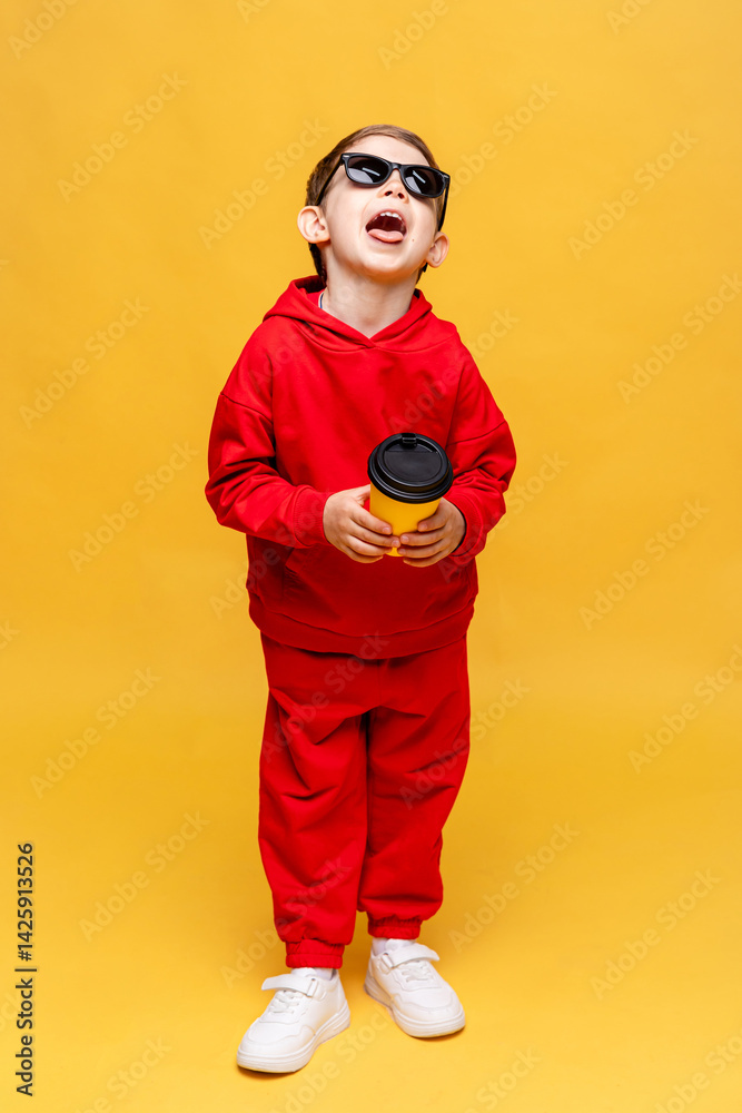 Cool little boy 3-4 years old in red track sport suit and black sunglasses holding yellow paper cup in hand on yellow background. Take away coffee concept. Copy space. High quality photo