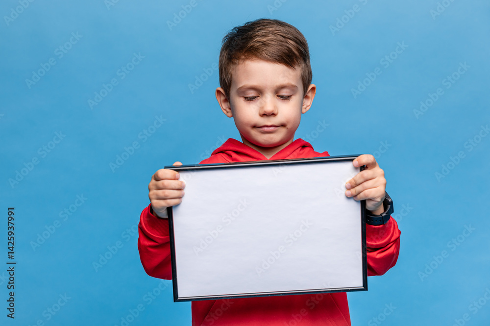 Portrait of happy laughing little boy holding white drawing board on blue background, young boy is holding a drawing board in his hands. Mock up. Copy space. High quality photo