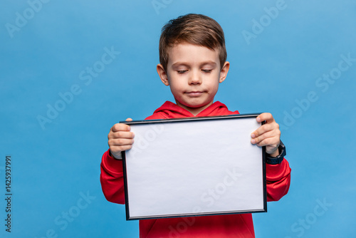 Obraz na płótnie Portrait of happy laughing little boy holding white drawing board on blue background, young boy is holding a drawing board in his hands. Mock up. Copy space. High quality photo w salonie