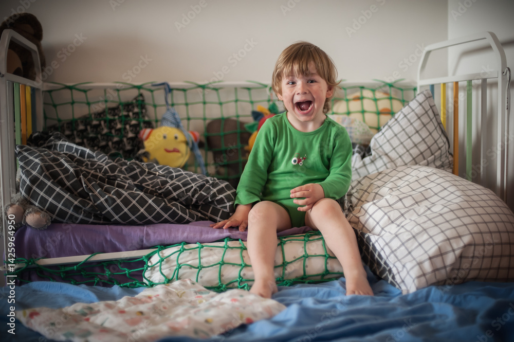 Naklejka premium A young child wearing a green shirt sits on a bed with various pillows and toys, laughing happily. The room is bright and inviting, creating a playful atmosphere.