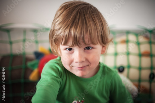 Portrait of a cheerful young child with blonde hair wearing a green shirt sits on a couch, surrounded by colorful plush toys and a playful atmosphere. The room is bright and inviting, showcasing joy.