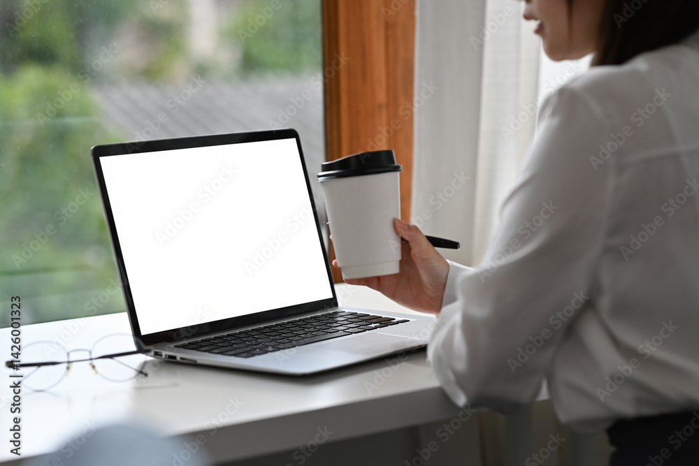 Close-up image of a businesswoman working remotely with a laptop and a takeaway coffee cup at a modern window desk. Blank screen laptop for mockup or design Close-up image of a businesswoman working remotely with a laptop and a takeaway coffee cup at a modern window desk. Blank screen laptop for mockup or design