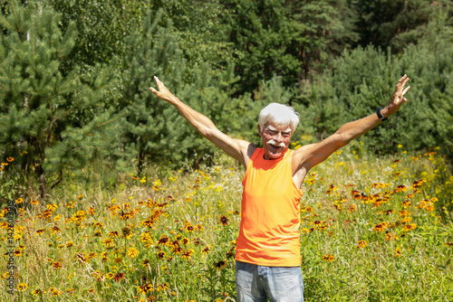 Positive smiley, funny casual, grey-haired handsome senior man looking straight into the camera lens while standing in a meadow among flowers - Eye contact - Portrait 