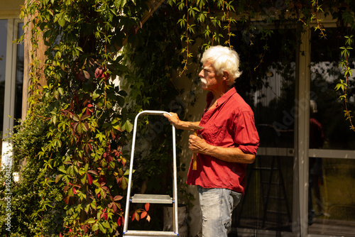 A handsome elderly gentleman in retirement doing small renovation work in his summer cottage. He is standing on the porch with a hammer in his hand next to a ladder.