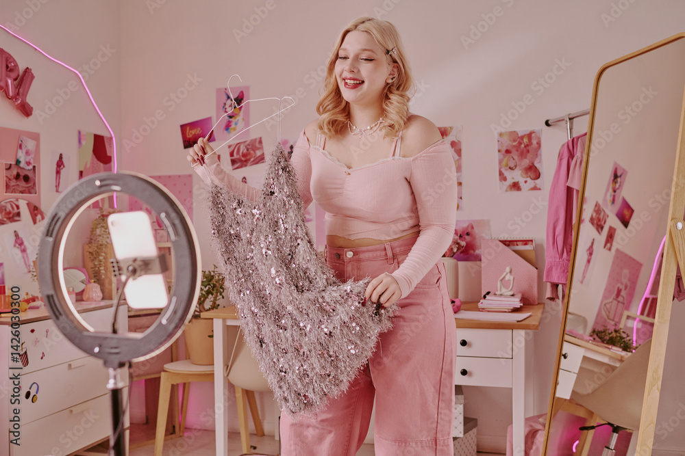 Obraz premium Professional teen blogger with doll vide holding dusty rose mini dress while recording purchase vlog at home