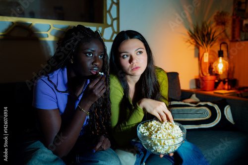 Two young women, an interracial lesbian couple, are sitting on a sofa at home, eating popcorn and watching a thriller movie on television